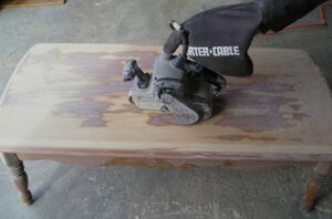 Using a belt sander to remove the existing finish and stain from a coffee table.