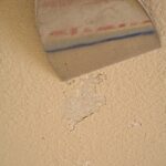 Using a stiff broad putty knife to remove loose paint on a wall.