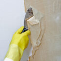 Using a broad putty knife to remove wallpaper and its backing paper.