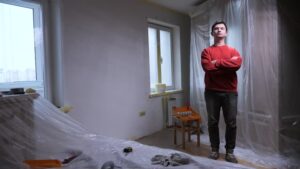 portrait of a man in his room where he paints the walls and does the renovation of the room with his own hands diy. Painter standing with arms crossed amidst drop cloths and painting tools in a freshly painted room during apartment renovation, showcasing a successful home improvement project